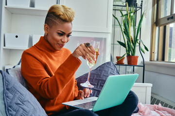 Smiling young woman flirting while drinking from champagne flute while video calling through laptop...