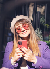Young happy woman in a purple beret coat and sunglasses sitting in a cafe with a smartphone in her hands