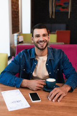 happy man sitting in restaurant with coffee cup near menu and smartphone with blank screen on table