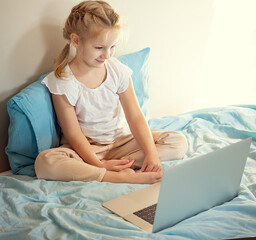 A cute girl sitting on the bed is watching something on a laptop. The concept of education.