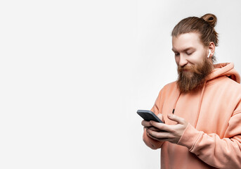 Scandinavian handsome happy man holding phone and smiling with wireless headphones in orange sweatshirt isolated on gray background. Guy with ginger hairstyle and beard. Modern digital technologies