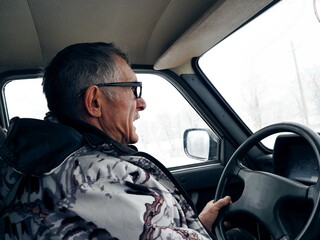 An elderly man in a winter jacket drives a Lada car in winter on a fishing trip. He is nervous and...