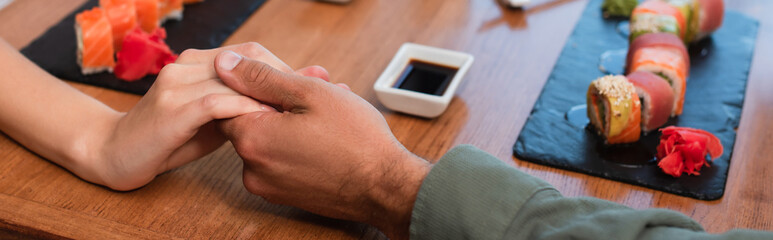 partial view of couple holding hands near sets of sushi rolls and bowl with soy sauce, banner