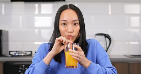 Portrait of a young Asian woman drinking orange juice. An attractive multiracial brunette girl is sitting in the kitchen at the table. Healthy eating.