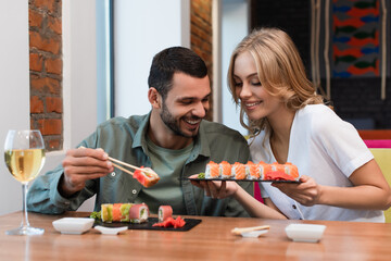 smiling woman showing delicious sushi rolls to boyfriend during dinner in restaurant