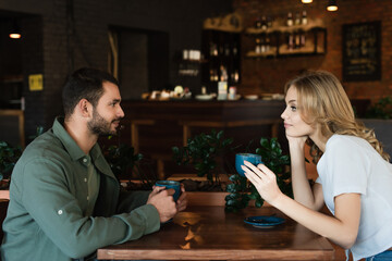 side view of young couple holding coffee cups and looking at each other in cafe