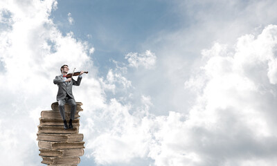 Handsome violinist in blue day sky on pile of books play his melody
