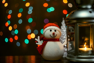 Christmas card: a snowman, a lantern with candles and blurry lights in the background.