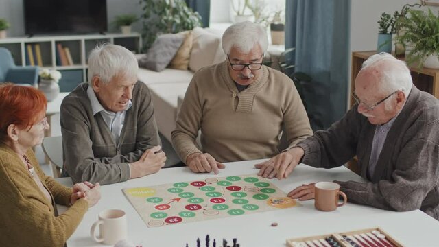 Medium shot of four Caucasian seniors playing board game together sitting at table in cozy nursing home