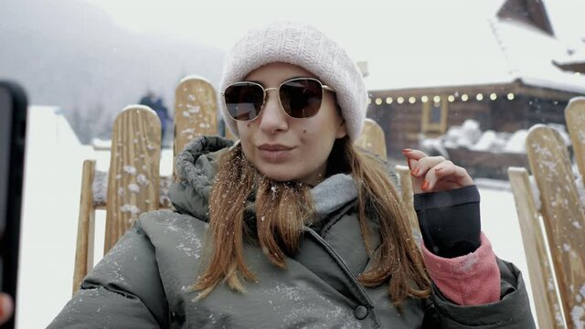 Fashionable young girl makes a selfie in sunglasses on a mobile phone under a snowfall in the mountains at a resort, insta blogger on vacation in winter