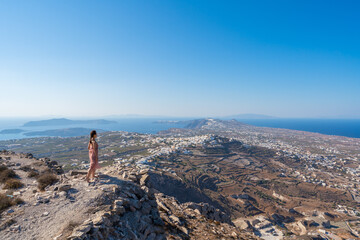 An Asian woman standing on the edge of a cliff overlooking the town of Pyrgos on Santorini island,...
