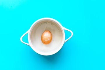 Ein braunes Ei mit aufgemalten Gesicht in einem weißen Kochtopf. Frühstück, Draufsicht, blauer...