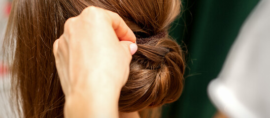 Hands of hairdresser making french twist hairstyle of an unrecognizable young brunette woman in a...