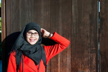 View of young Asian muslim woman wearing black hijab and red coat leaning on ancient wooden gate in Kyoto, Japan. Excited and happy expression. Face with bright sunlight.
