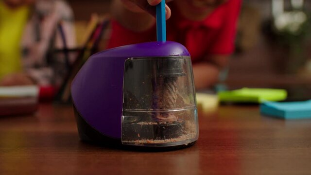 Close-up of child hand sticking pencil into electric pencil sharpener and sharpening while African American kids drawing picture in domestic interior.