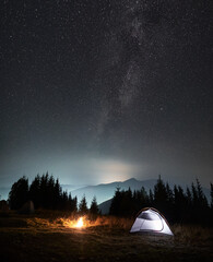 Magnificent view of night starry sky and Milky way over grassy hill with illuminated camp tent and campfire. Concept of hiking, night camping and astronomy.