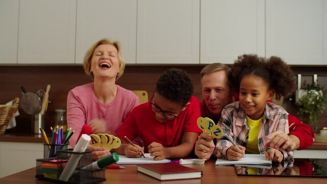 Caring attractive middle aged grandparents developing lovely primary school multiethnic grandchildren, teaching english alphabet together while multi generation family studying indoors.