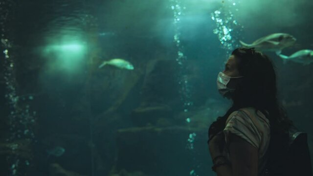 The girl watches the aquatic animals in the aquarium with mask on her face during Covid19 pandemic.