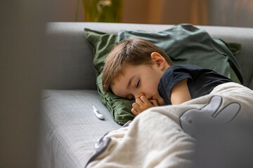 Unhealthy little boy child measure high temperature with thermometer sleep in bed relax at home in...