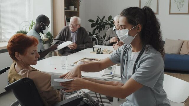 Medium shot of young female nurse in face mask taking blood pressure of senior woman in wheelchair at nursing home while her African-American male colleague talking to senior men in background