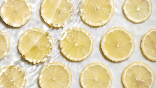 Waves and splashes on water surface over lemon slices arranged in lines on white background | skincare background, face wash commercial