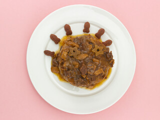 Canned meat with sauce decorated with dry pet foods on white plate isolated on pink background. Cheerful face made of various types of pet foods. Top view