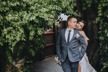 Fototapeta na wymiar Stylish groom and beautiful bride tenderly embrace against the background of a wall with green foliage, ivy leaves. Wedding portrait of smiling, happy newlyweds.