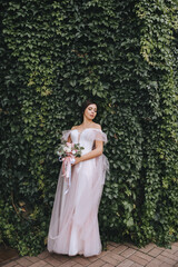 A beautiful brunette bride in a white dress stands against a wall of green ivy foliage. Wedding portrait.