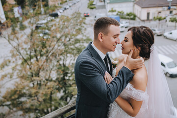 Stylish groom and beautiful bride in a white dress are standing, hugging on the balcony, veranda against the backdrop of the city with buildings. Wedding portrait of young newlyweds.