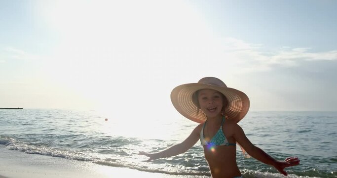 A child with a large straw hat on his head is resting on the seashore in the summer, turns to face the video camera and smiles in surprise.