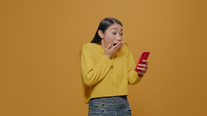 Amazed woman reading good news on smartphone, standing over yellow background. Asian person feeling cheerful after having surprised reaction while she uses mobile phone and social media.