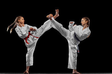 Two young girls, teens, taekwondo athletes training together isolated over dark background. Concept...
