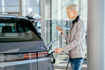 Eco electric car concept. Blond female waiting electric car to charge. Business woman with the...