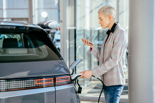 Eco Electric Car Concept. Blond Female Waiting Electric Car To Charge. Business Woman With The Charging Cable Near The Electic Vehice, Hybrid Cars. Concept Of Buying Electric Vehicle.