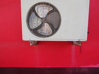 The old air cooled condensers  hangs on a bright red painted plaster wall. The air compressor fans are old and discolored due to outdoor installation and exposure to sunlight.
