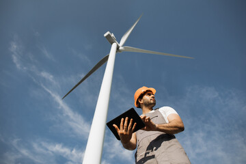 Indian engineer in orange helmet and grey overalls standing among windmill farm with modern tablet...