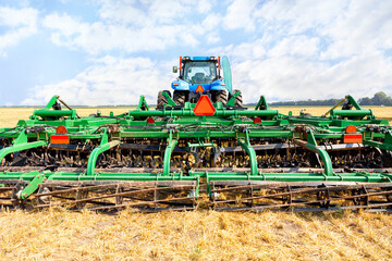Fototapeta premium An agricultural tractor with a trailed harrow against the background of a harvested wheat field on a warm autumn day. Central composition.