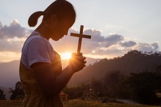 Silhouette of young girl praying to the GOD while holding a crucifix symbol with bright sunbeam on the mountain