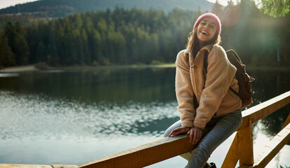 Pleased laughing woman sits on pier near mountain lake. Tourist woman enjoying the day outdoors in nature landscape - 477467516