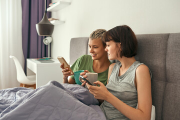 Lesbians with tea using smartphones on bed at home