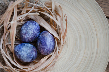 Easter eggs in trendy purple very peri color of the year 2022. Chicken eggs in a straw nest on a wooden plate. Eco-friendly natural coloring of products. Zero waste concept. Copy space.