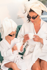 Mother with child doing beauty treatment together. Happy family mother and child daughter in towel and bathrobe are sitting on the sofa and drinking tea
