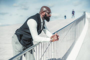 A bald black man in elegant attire leaning on the chrome bridge railing on a sunny day. Bearded gentleman with glasses on the footbridge looking ahead. Businessman on the bridge looking at the street