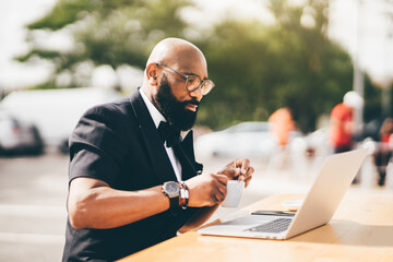 Selective focus on a stylish bearded gentleman sitting outdoors, using a notebook, and holding a cup. A black man in glasses at a cafe table, looking at his notebook screen while stirring his coffee - obrazy, fototapety, plakaty