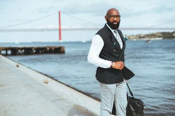 A businessman holding a messenger bag near a river, a red suspension bridge in a blurred background. A bald bearded black guy in a suit standing on the pier looking away, a copy space area on the left
