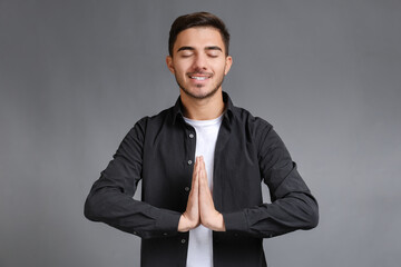 Bearded smiling young man with closed eyes wearing black shirt keeping hands namaste gesture and meditating on gray background. Yoga exercise breath technique reduce stress and increase concentration