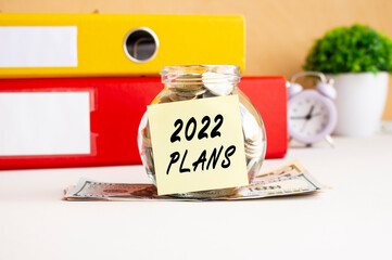 A glass jar with coins stands on a stack of dollar bills on the table. The jar has a sticker with the text 2022 PLANS.