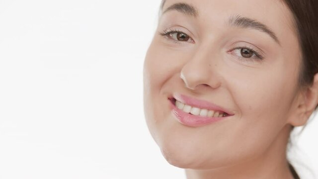 Horizontal big close-up of beautiful Caucasian young plus size woman with long dark hair strokes her cheek and looks at the camera smiling wide on white background | Skin moisturizing concept