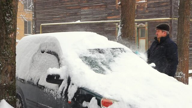 A man cleans the roof of a car from snow with a brush. Cloudy cool winter day, fresh snow is falling from the sky. Auto like a snowdrift. Climate change, city, snowstorm. UHD 4K.