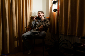 A 20 something man sitting in an arm chair talking on his mobile phone on a dark winter evening with a lamp lit. Life and after dark concept